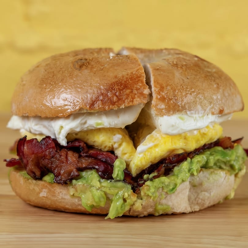 A bagel sandwich with scrambled eggs, bacon, cream cheese, and avocado on a wooden board.