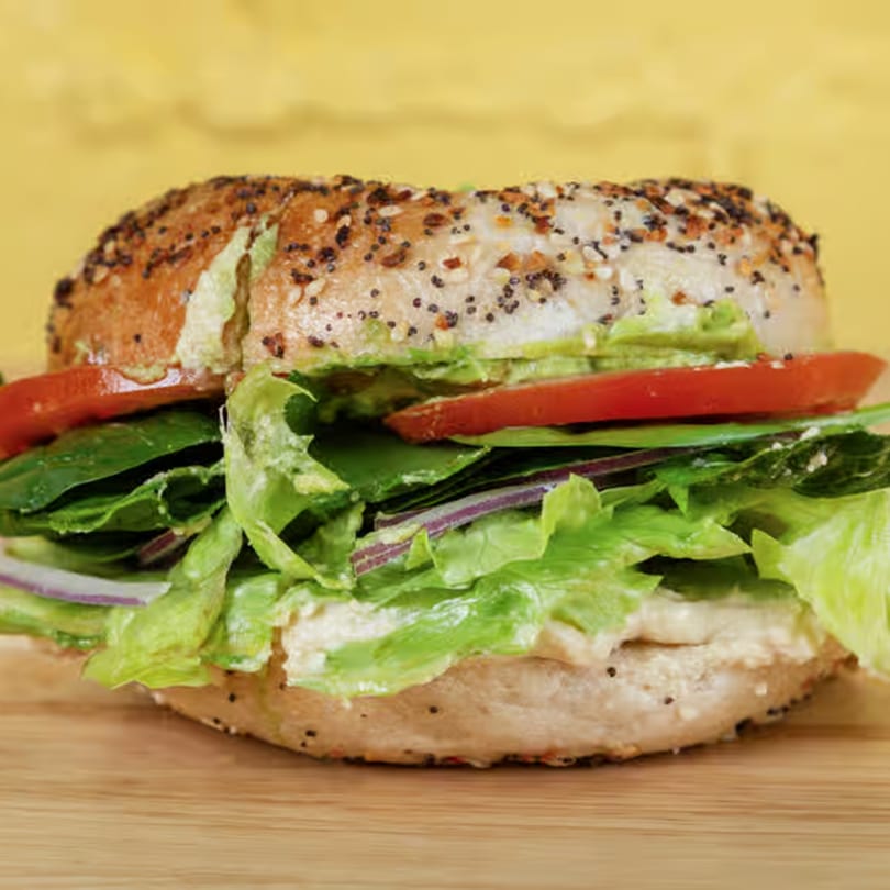 Everything bagel sandwich with hummus, lettuce, tomato, and red onion on a wooden board.